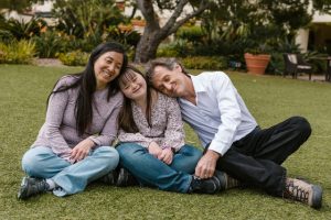 A mother and father sitting with their daughter (person with Down syndrome) on a grassy lawn.
