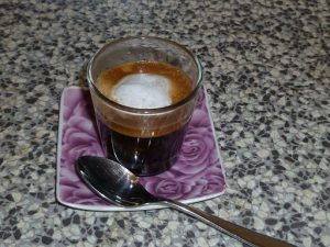 Coffee drink with dollop of white foam all in a clear glass. There is a spoon resting on the saucer