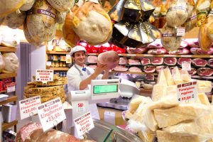 Man holding mortadella in a market surrounded by meats and cheeses