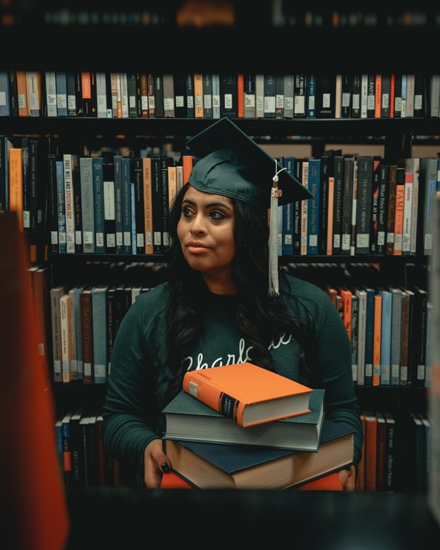 Person with a graduation cap on holding a stack of books in a library