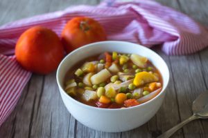 Soup with many vegetables on a table with two tomatoes, a napkin and spoon.