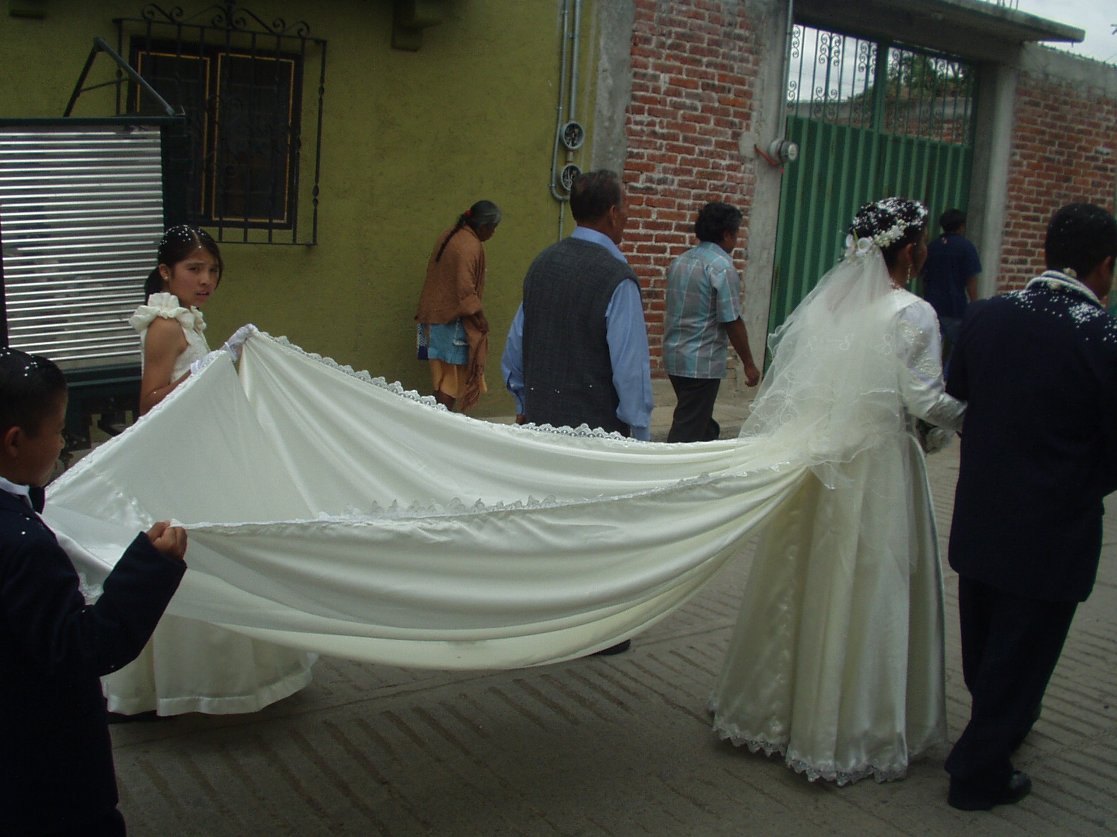 Wedding procession where a bride and groom walk toward the right of frame with linked arms. The bride wears a white dress with a long train carried by two children walking behind her. A few others walk alongside the bride and groom.