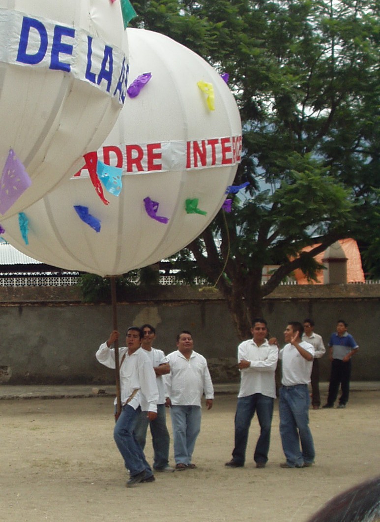 Four men in jeans and white shirts, one of whom carries a large wood and cloth sphere with some colorful details on a stick