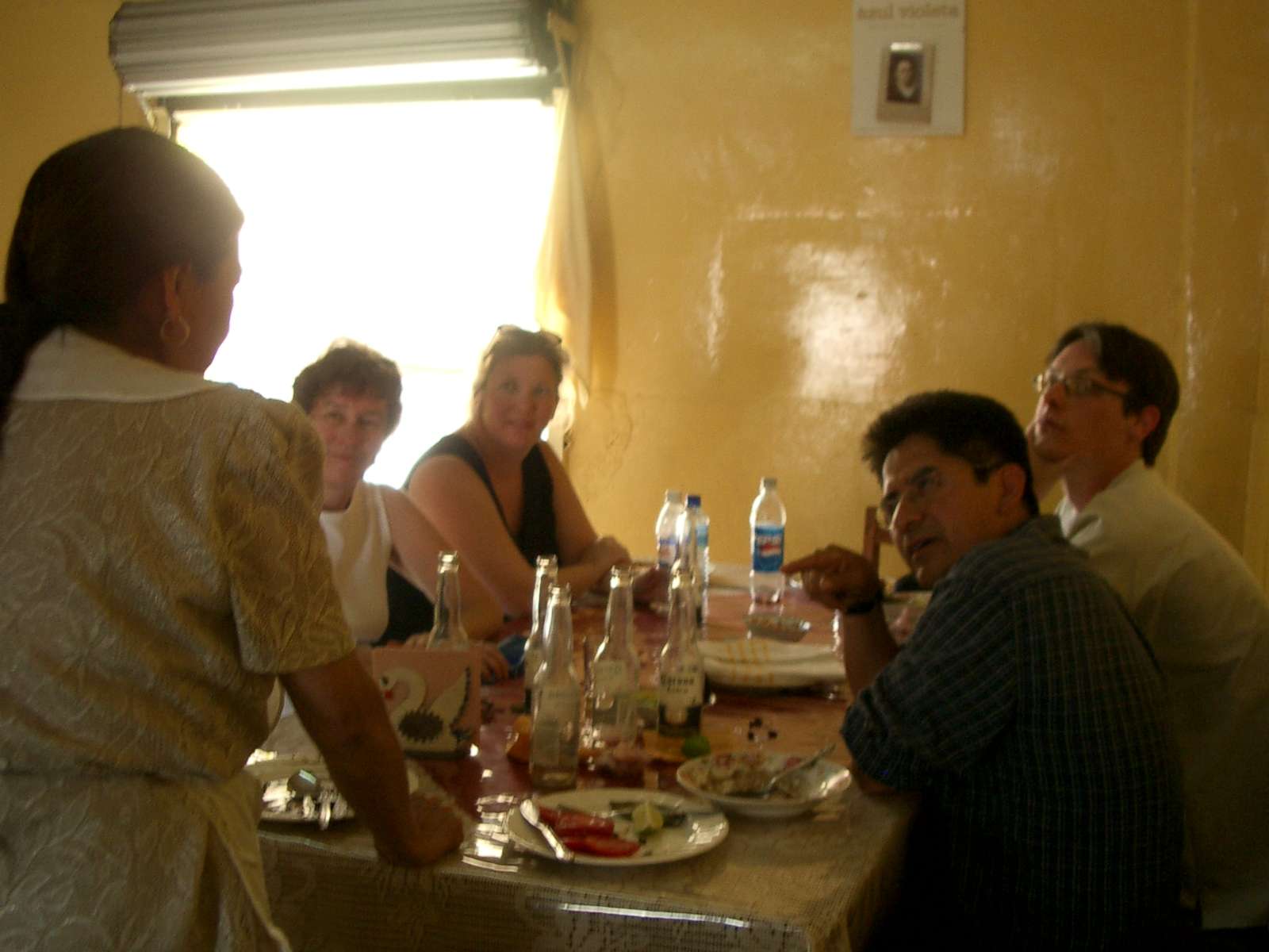 Four people sitting at a table with food and drinks, facing a standing woman whose back is to the camera.