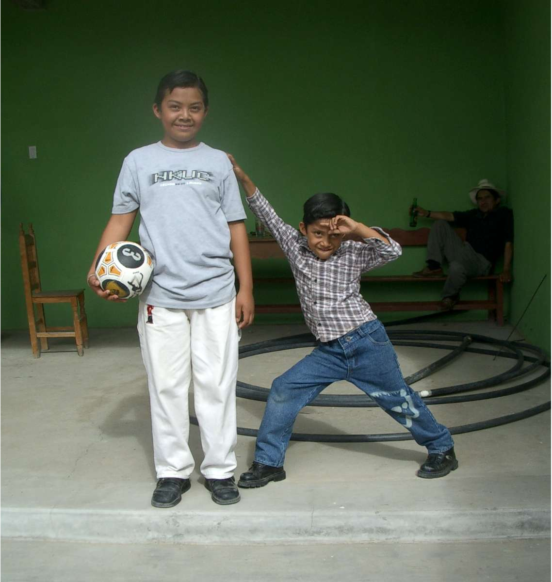 Two children, the taller one to the left standing and holding a soccer ball, and the shorter one to the right striking a pose and reaching his arm up to touch the taller child's shoulder.